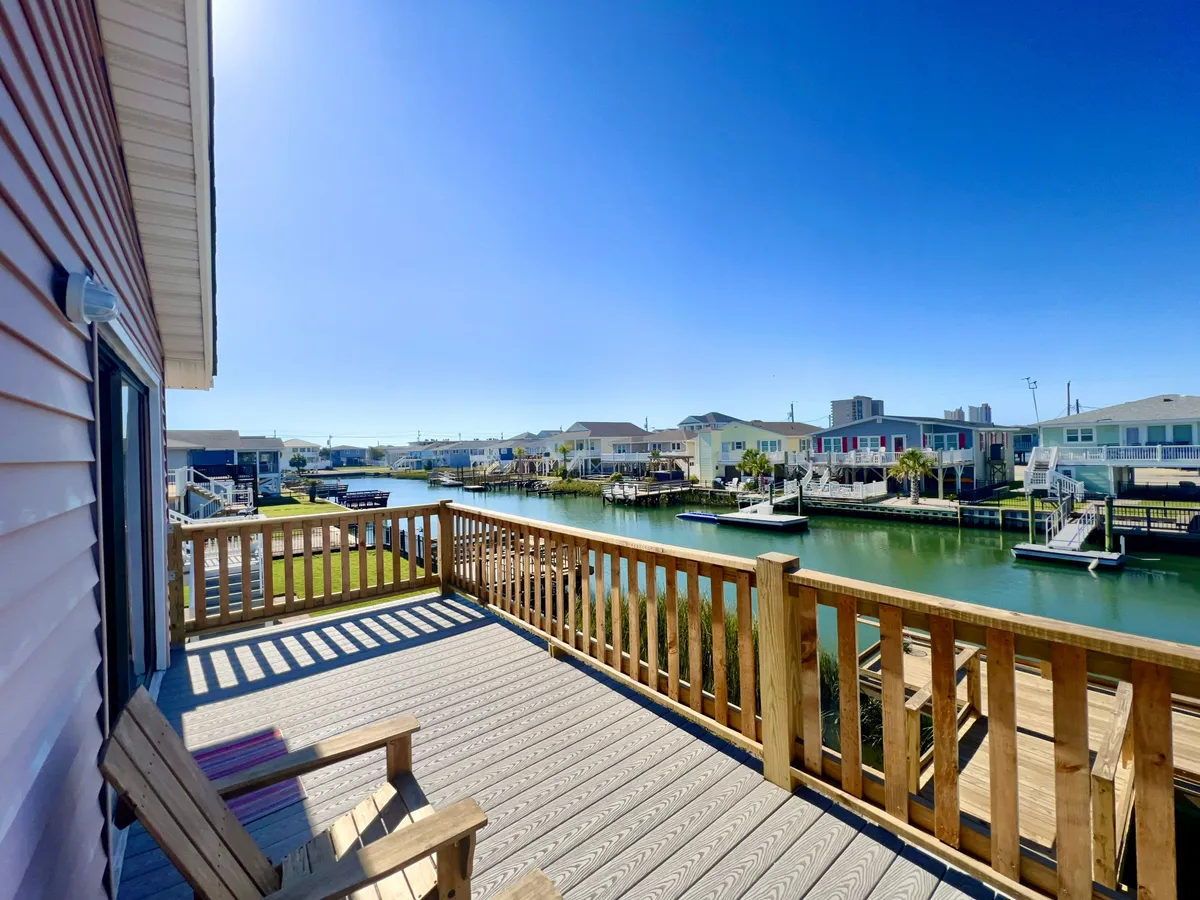 An inviting deck area overlooking a picturesque water canal, with ample space for groups to relax, fish, or host a summer BBQ. The deck is surrounded by other homes and lush greenery, showcasing the communal and social nature of the neighborhood.