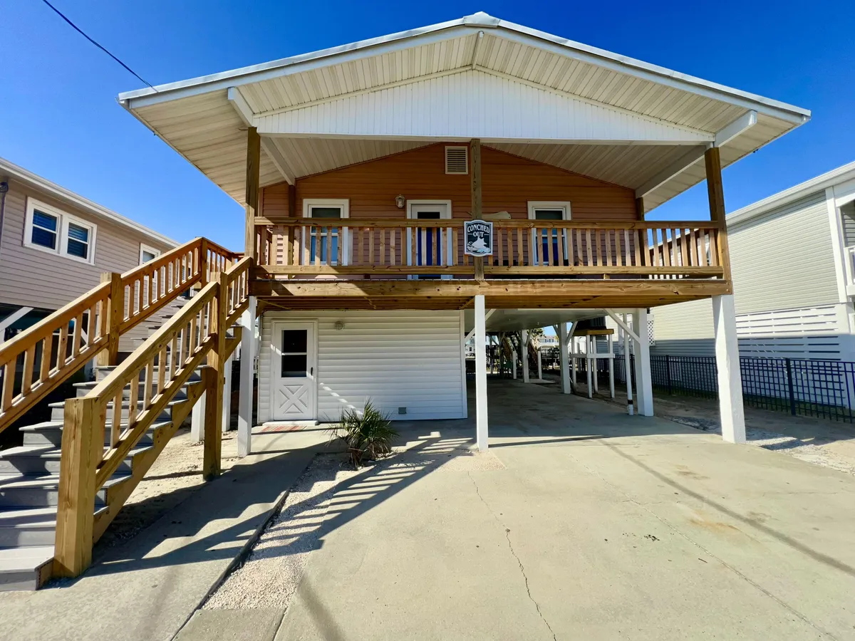 The image features the exterior of a spacious single-family house with a prominent wooden staircase leading up to a wide balcony. The balcony is perfect for gatherings and overlooks the serene waterfront, with sunset hues casting a warm glow. The home’s name, 'Conched Out,' is displayed on a cheerful sign, and there is plenty of space for outdoor dining and parking. The atmosphere is welcoming, hinting at relaxation and fun with loved ones.