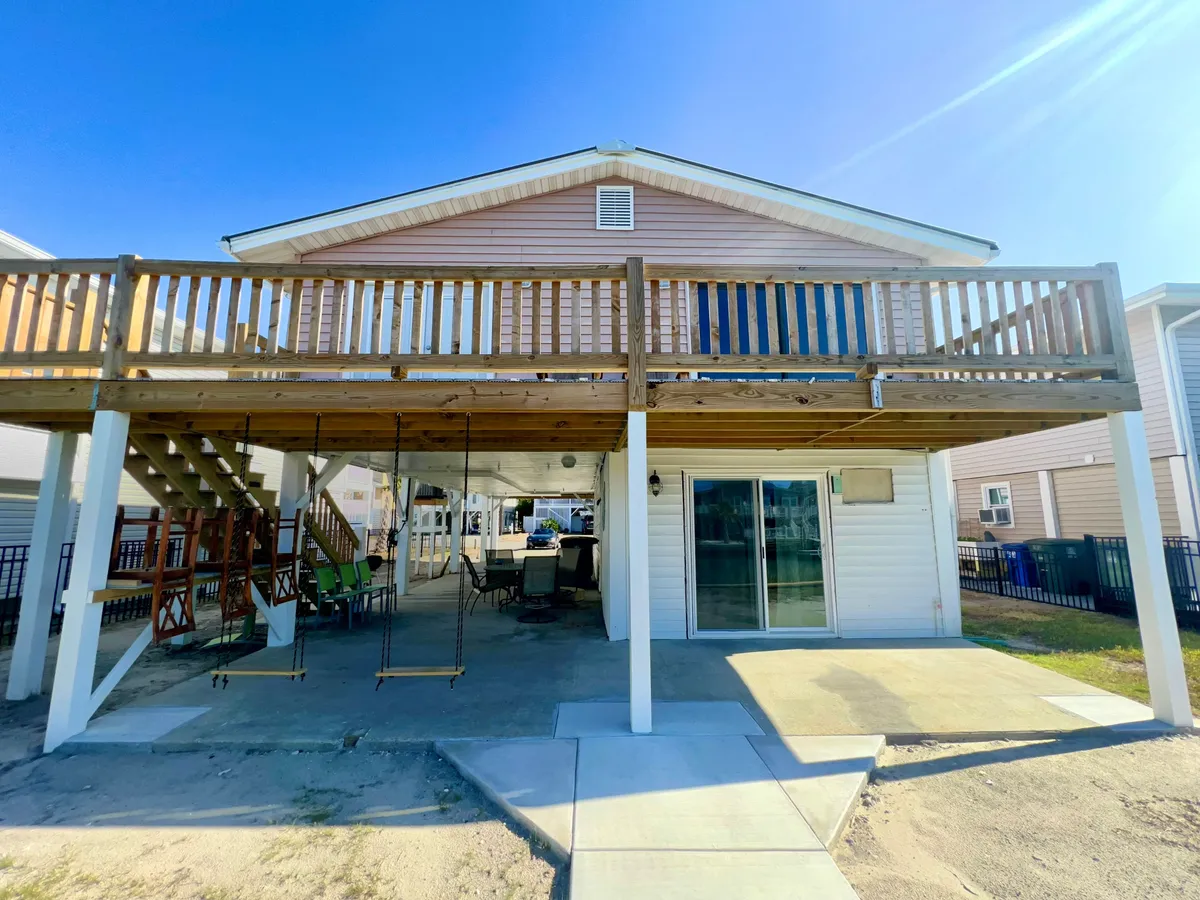 An inviting exterior shot showing a spacious deck with a railing, a swing set for children, a large patio area, and a sliding glass door leading into the house. The scene is bright and cheerful, ideal for both family relaxation and group gatherings.