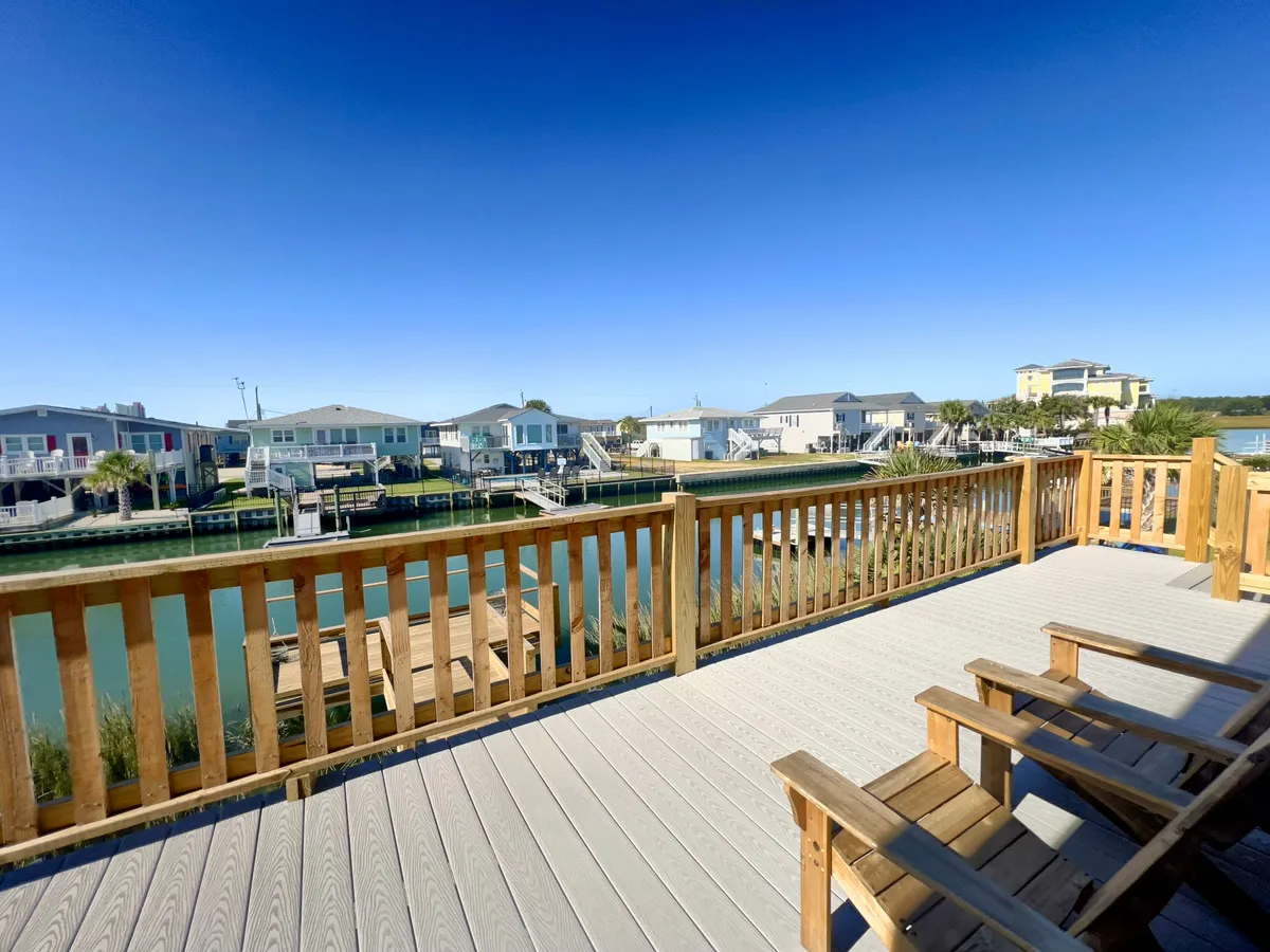 A serene view from another angle of the wooden deck, this time emphasizing the open blue sky, calm waterway, and proximity to neighboring houses. The deck is styled for relaxation and interaction, with opportunities for fishing, socializing, and watching boats pass by.