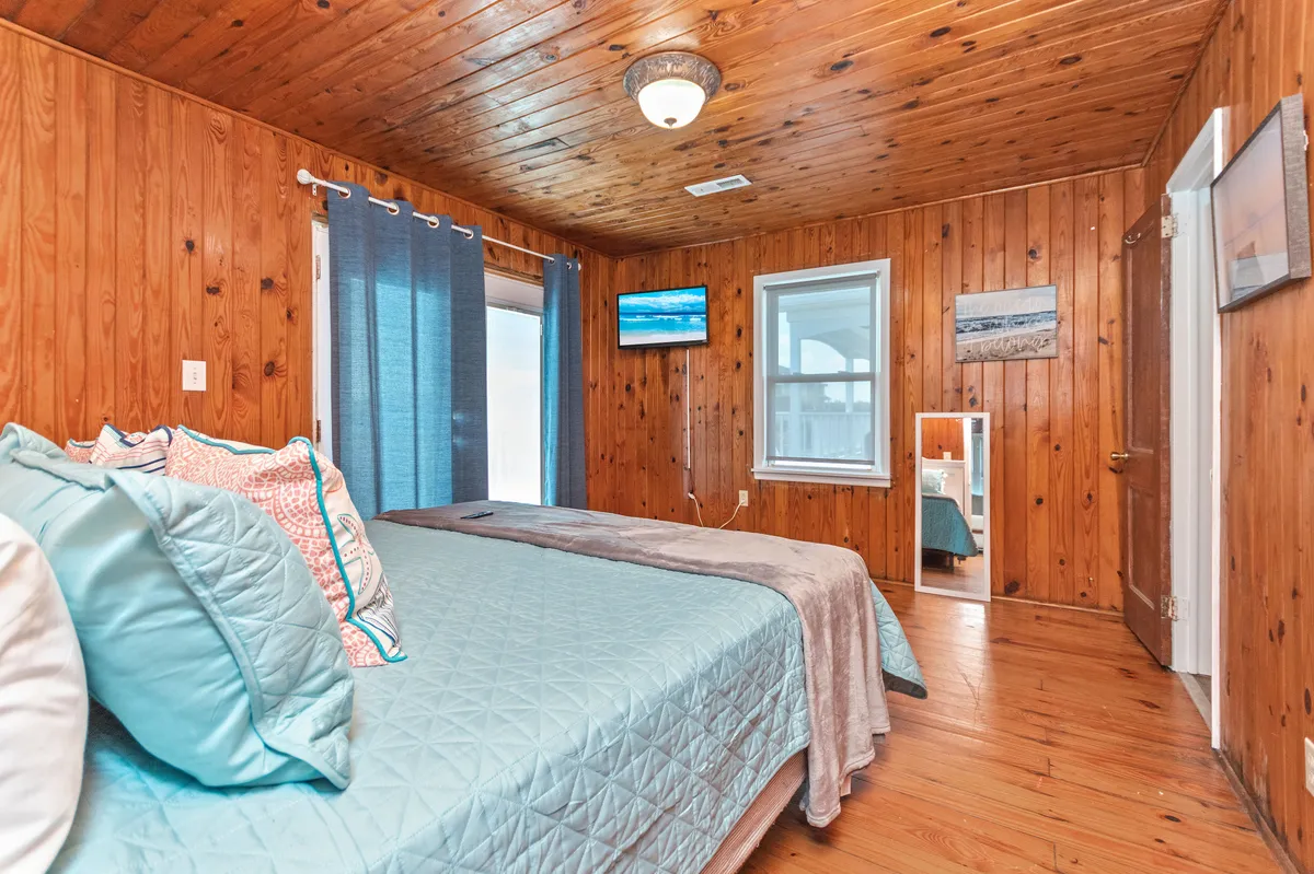 This tranquil bedroom features a bed with teal bedding, wooden walls, a wall-mounted TV, and blue curtains. The atmosphere is serene and private, suggesting a comfortable space for solitude or relaxation—an excellent representation of a downstairs suite.