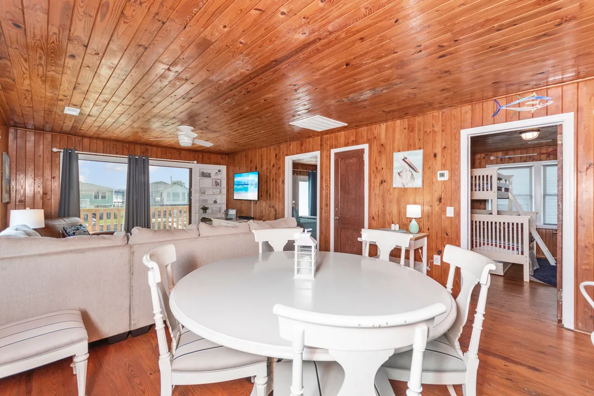 A casual interior scene featuring a round dining table, comfortable seating area, large windows, and a TV. The open layout allows for seamless transitions between dining, relaxing, and group entertainment.