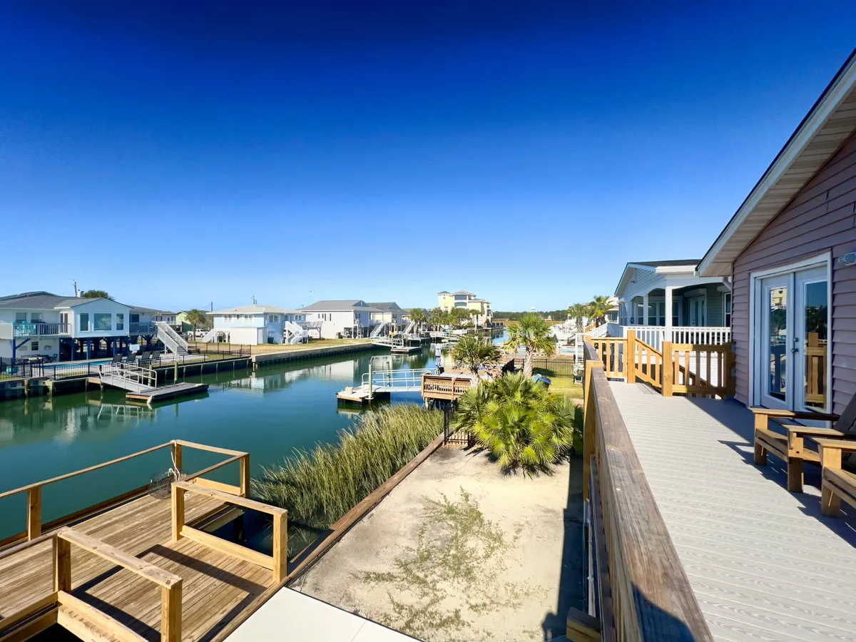 A wide view of the wooden deck overlooking a tranquil waterway, with palm trees and neighboring houses in the distance. The setting features space for relaxing, gathering, and enjoying the scenery, making it ideal for outdoor social activities. Boats can be seen on the water, reinforcing the access to water adventures.