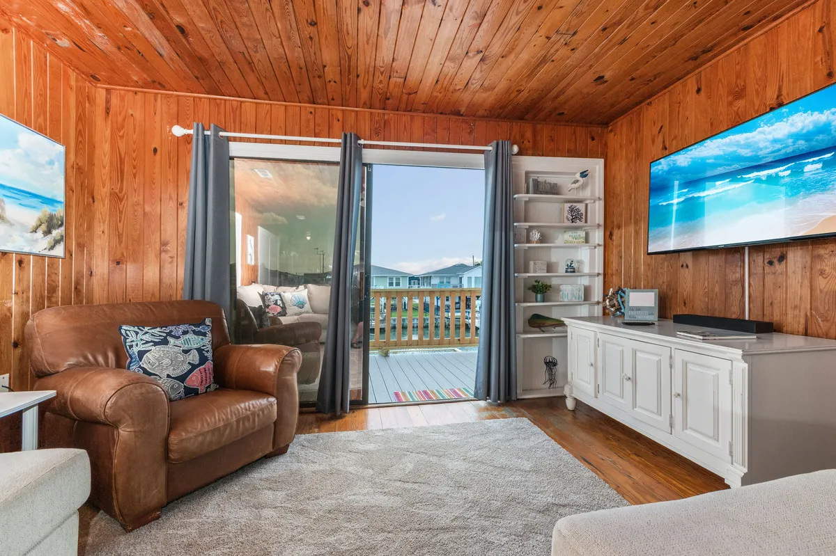 A cozy living area with wooden walls, a sliding glass door leading to an outdoor deck, a comfortable armchair, TV, and decorative shelving. The soft light hints at sunrise or sunset, and the arrangement is ideal for intimate movie nights or social gatherings inside with a seamless connection to the outdoors.