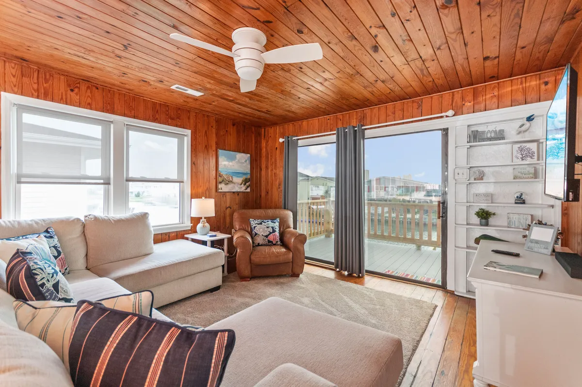 A spacious living room with a comfortable sectional sofa, wooden ceiling, large windows, a sliding glass door, and decorative items on a shelf. Natural light floods the space, emphasizing the inviting atmosphere for group relaxation, reading, or enjoying the view.