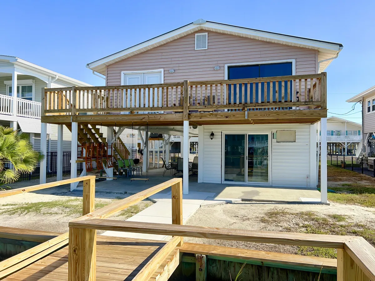A wide outdoor deck with ample seating, large glass doors connecting the interior, and a palm tree for shade. The space is perfect for family gatherings, outdoor dining, and relaxation, all while enjoying views of the water. The seamless transition between indoor and outdoor spaces is apparent, reinforcing the home's coastal, open-concept living.