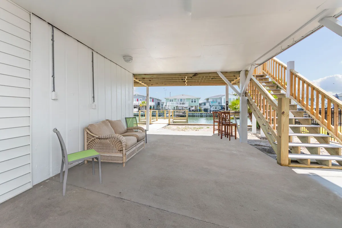 This image features a cozy wicker seating area with a bar table and stools, set on an exterior deck overlooking the water. The area seamlessly blends indoor comfort with an outdoor setting, offering an inviting space to socialize, relax with a drink, or watch boats glide by. The wooden staircase and open view reinforce the indoor-outdoor flow and the casual, beachy atmosphere.