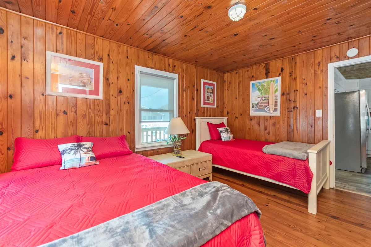The image displays a welcoming bedroom with two beds featuring red bedding, wooden walls and ceiling, decorative wall artwork, and a lamp on a nightstand. Natural light streams in, creating a cozy and rejuvenating space ideal for relaxation and family togetherness.