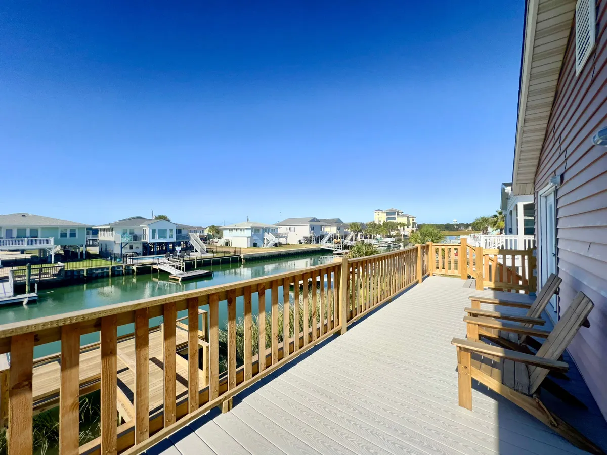 A spacious wooden deck overlooking a tranquil waterway, with neighboring houses visible across the canal. The deck offers plenty of space for outdoor gatherings, with room for seating, grilling, and enjoying waterfront views under the open sky.