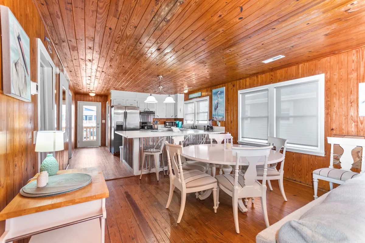 A bright dining area with a large dining table, kitchen island, wooden ceiling, and expansive windows that let in sunlight. The setup encourages shared meals and social gatherings, with enough space for families and friends to dine comfortably.