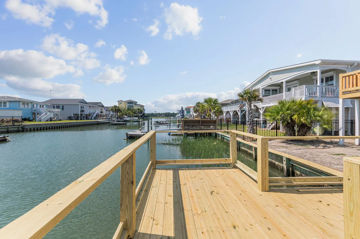 A wooden deck overlooking a serene waterway, bordered by palm trees and neighboring houses. The deck is a tranquil spot ideal for relaxing or enjoying the sunrise with pets by your side.