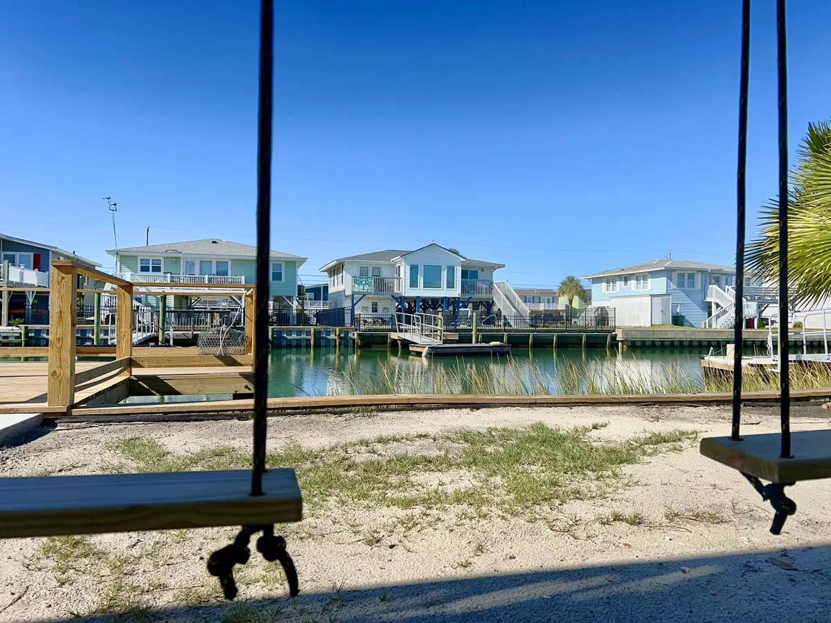 A tranquil exterior shot showing a swing set positioned near a waterfront dock and surrounded by neighboring homes. The area is open, grassy, and welcoming, making it ideal for children and pets to play safely by the water. The swing, dock, and spacious yard reinforce the idea of outdoor recreation and pet-friendly amenities.