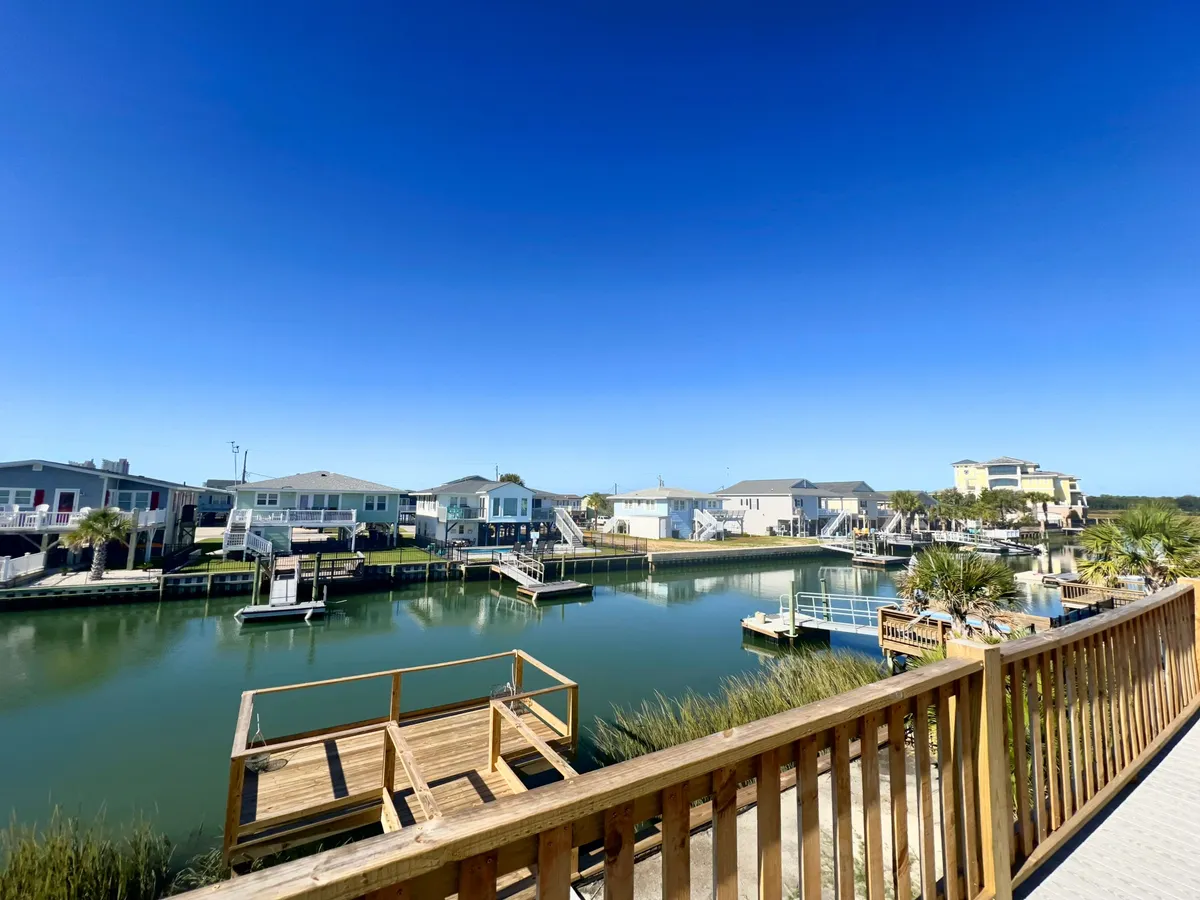 A serene waterfront view with calm water, a wooden dock extending out, green grass, and nearby homes. The scene is peaceful, perfect for watching sunsets, fishing, boating, or simply relaxing by the water with loved ones.