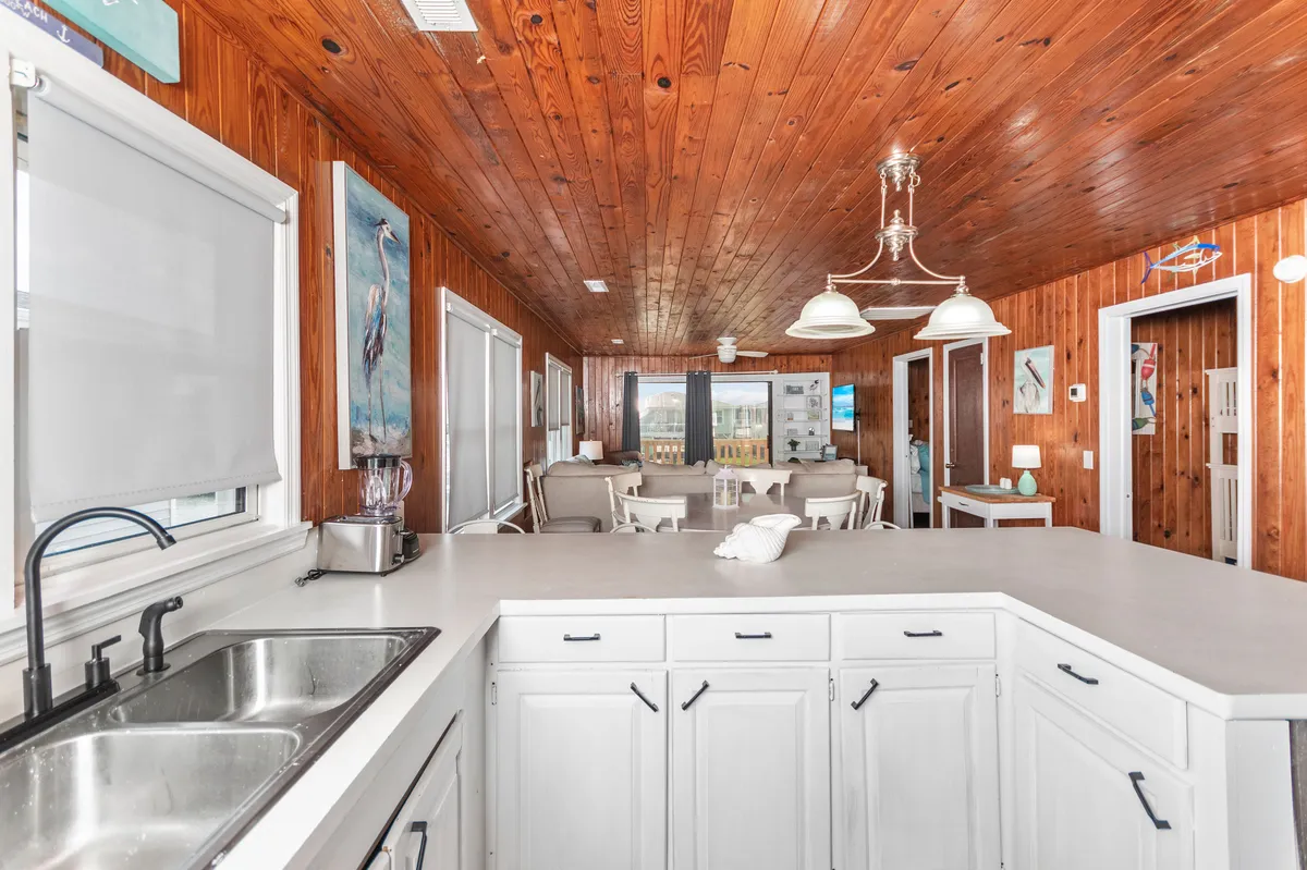 Here, the kitchen sink and ample counter area are visible, with a view into the adjacent living area. The wooden ceiling and wall art give the space warmth, while the clear sightlines suggest the chef can interact with others or enjoy the water views mentioned in the narrative.